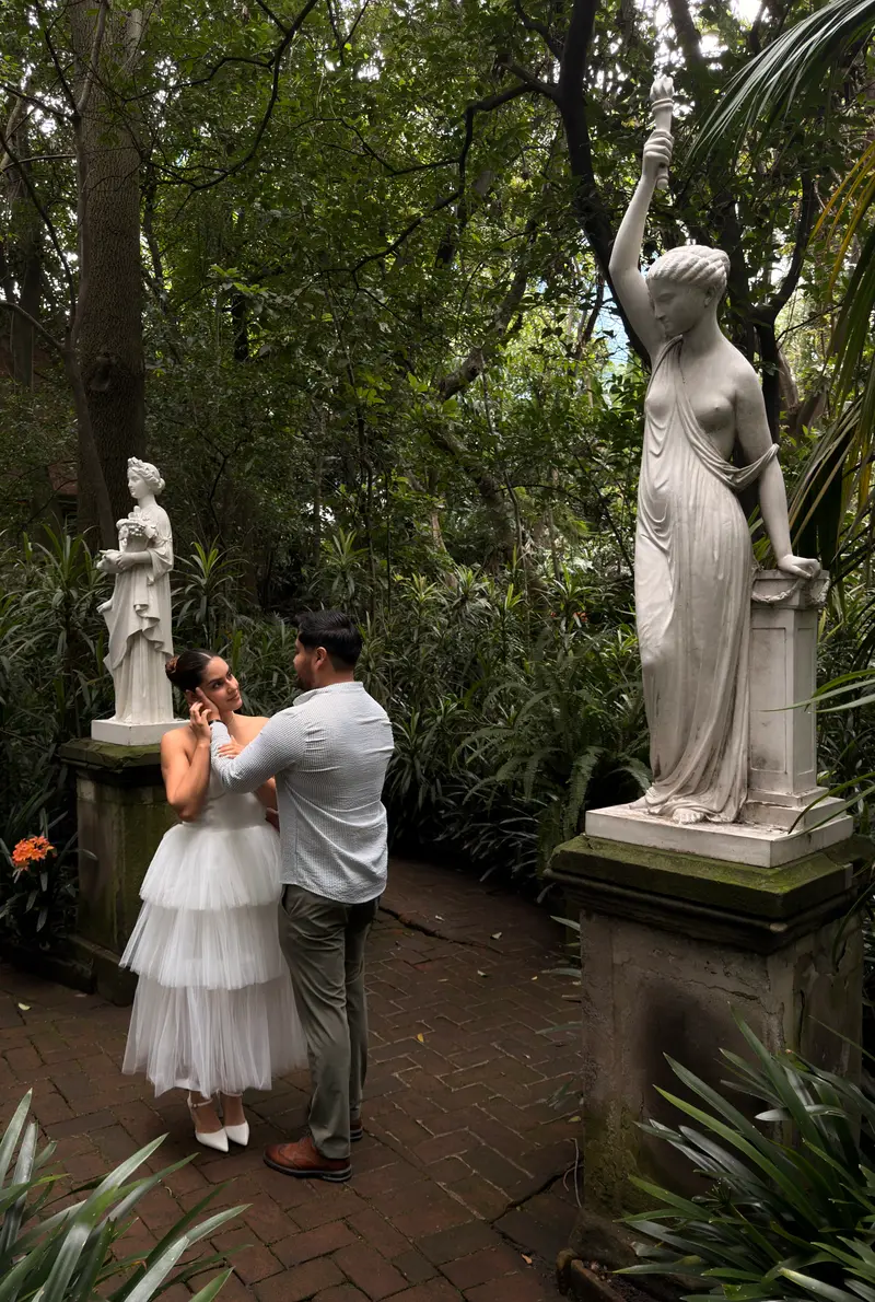 Karyme y Josué frente a frente en el sendero del jardín, junto a una estatua de mármol